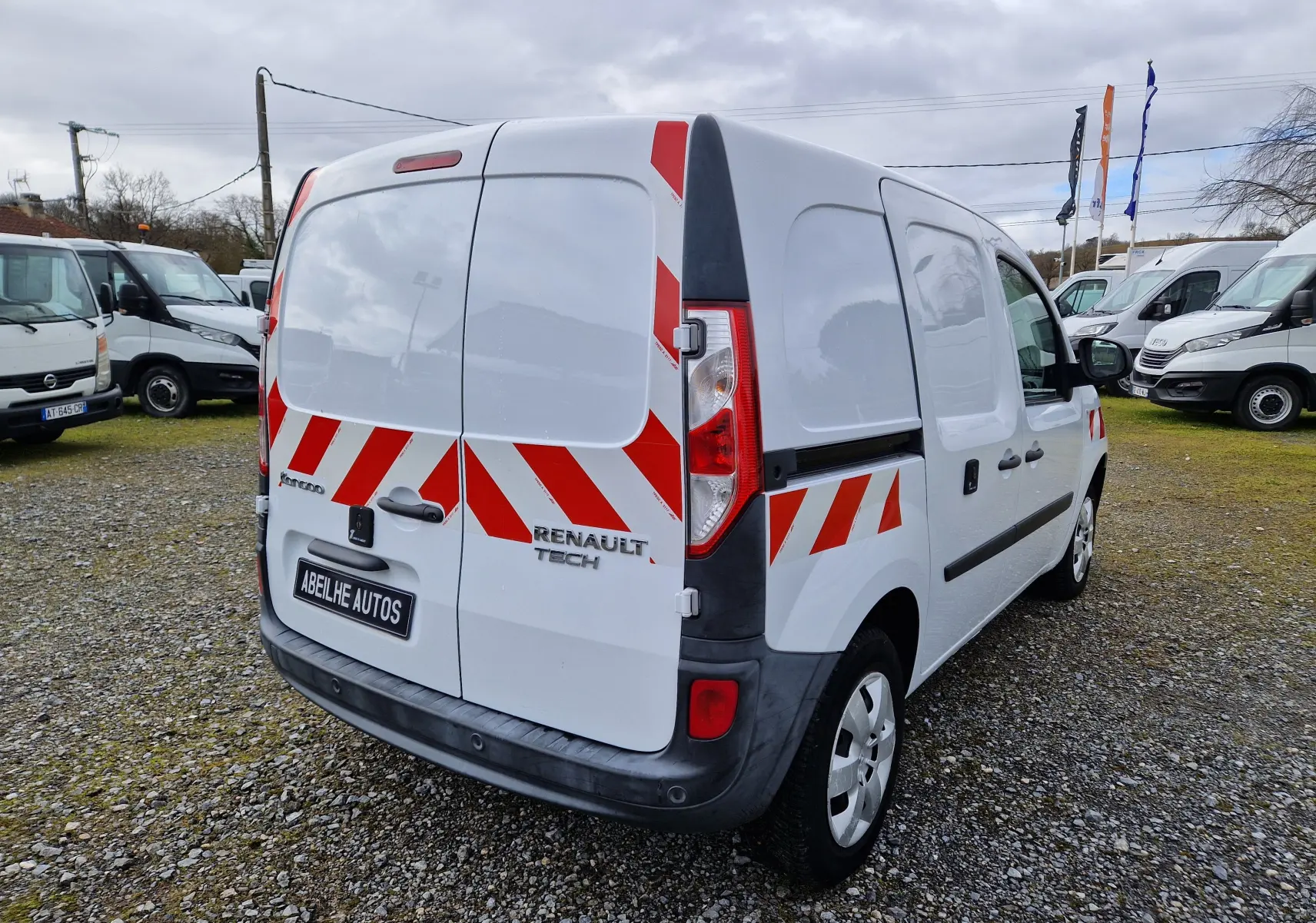 Vue 3/4 arrière droite d’un Renault Kangoo blanc avec bandes rouges réfléchissantes, stationné sur un sol gravillonné.