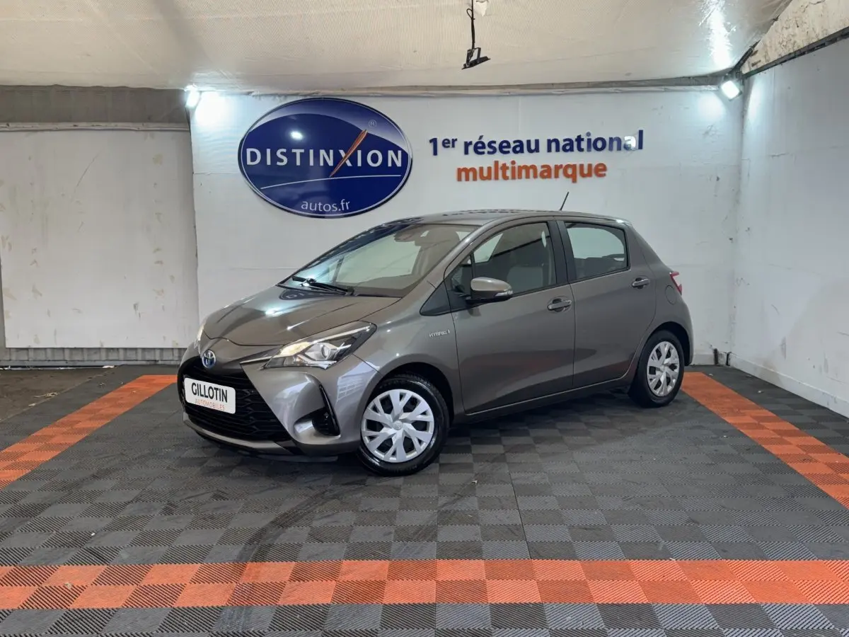 Toyota Yaris gris foncé vue 3/4 avant droit dans un showroom avec sol à damier noir et orange.