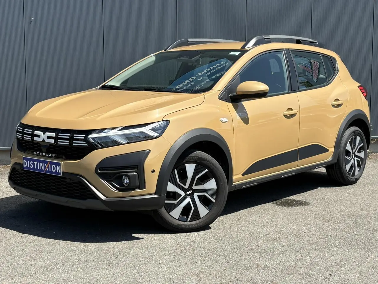 Vue 3/4 avant droite d'une Dacia Sandero Stepway beige Safari avec barres de toit et jantes spécifiques.