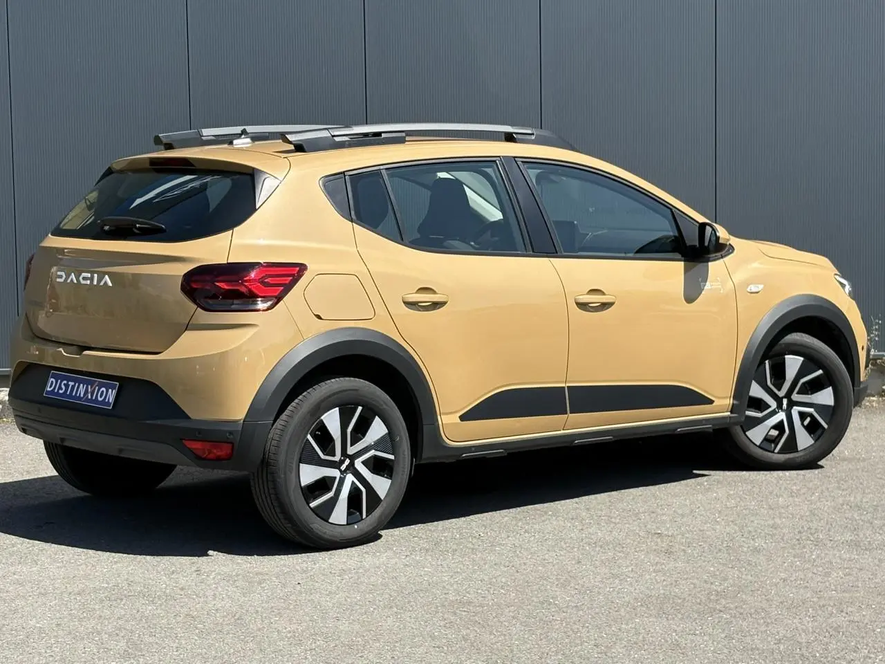 Dacia Sandero Stepway beige Safari vue 3/4 arrière droit avec barres de toit et jantes noires et argentées.