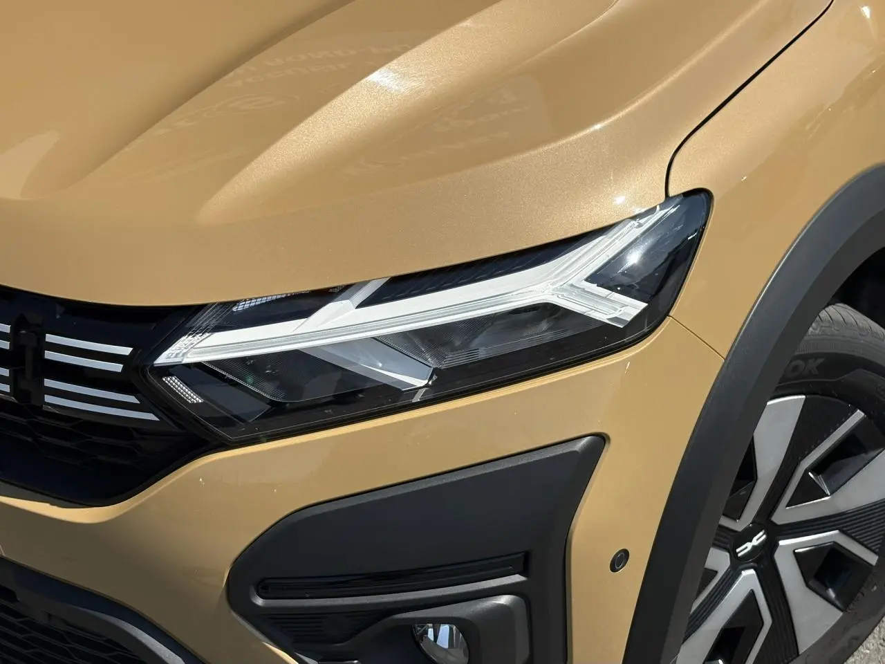 Gros plan sur l'avant droit beige Safari du Dacia Sandero Stepway 2025, mettant en valeur le phare LED et la roue avec enjoliveur.