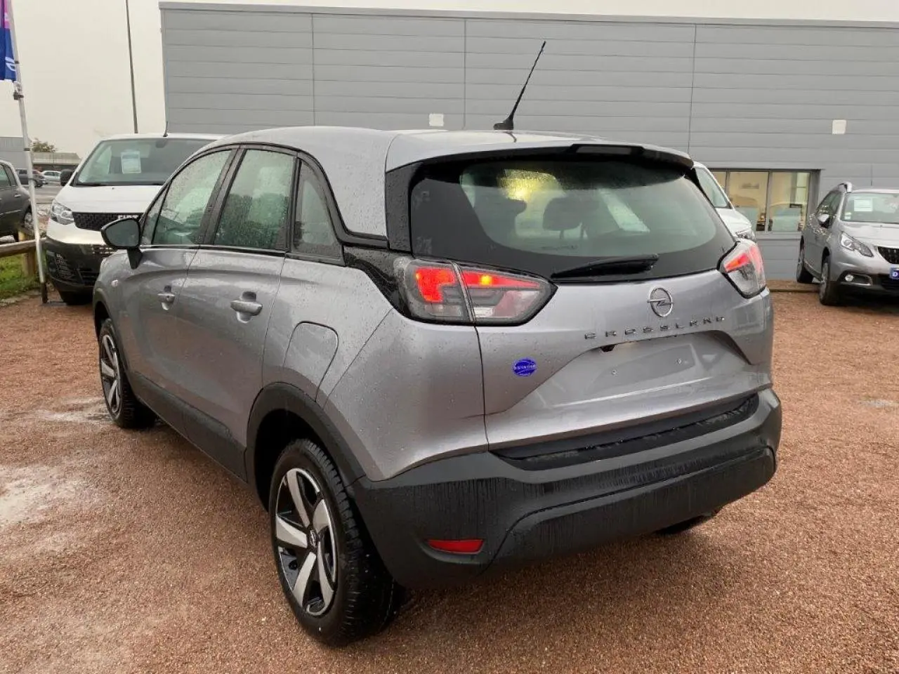 Vue 3/4 arrière droite d'un Opel Crossland gris métallisé avec feux arrière allumés sur parking humide.