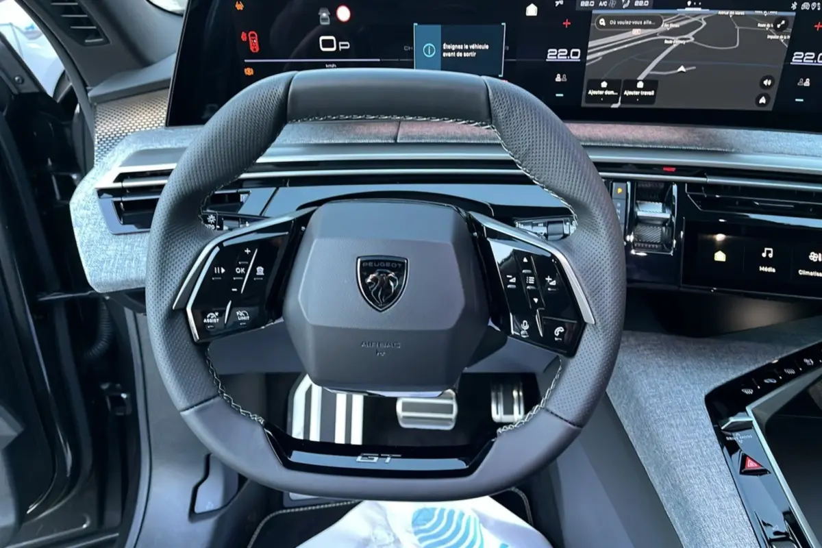 Vue rapprochée du volant cuir multifonctions Peugeot 5008 Hybrid 145 GT Alcantara avec tableau de bord numérique et console grise.
