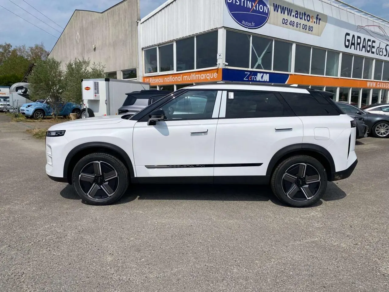 Vue de profil côté gauche du SUV JAECOO J7 blanc khaki avec toit noir et jantes modernes, stationné devant un garage.