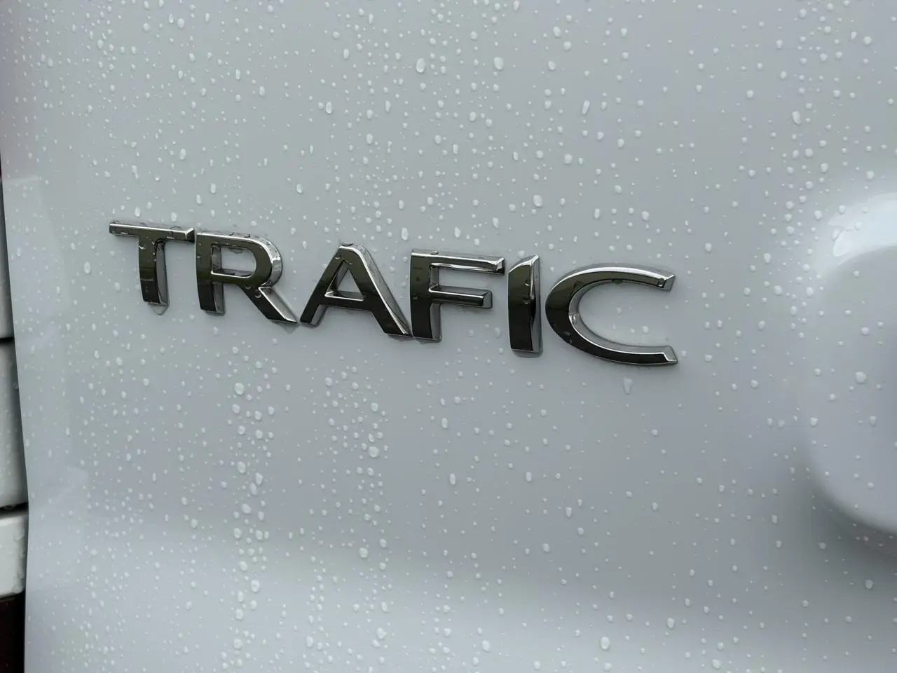 Gros plan sur l’inscription TRAFIC en chrome sur la porte arrière d’un Renault Trafic blanc avec gouttes d’eau.