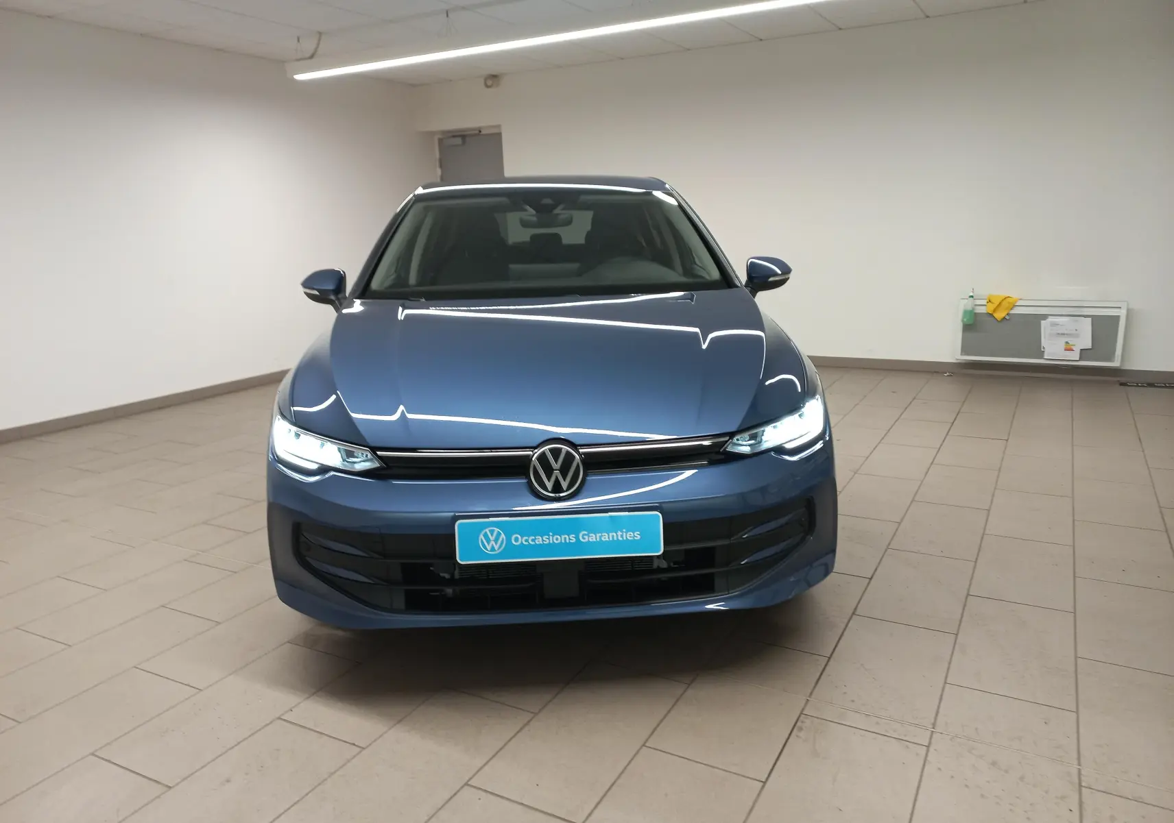 Vue avant d'une Volkswagen Golf 1.5 TSI Bleu Anemone avec phares allumés et plaque "Occasions Garantis" dans un showroom.