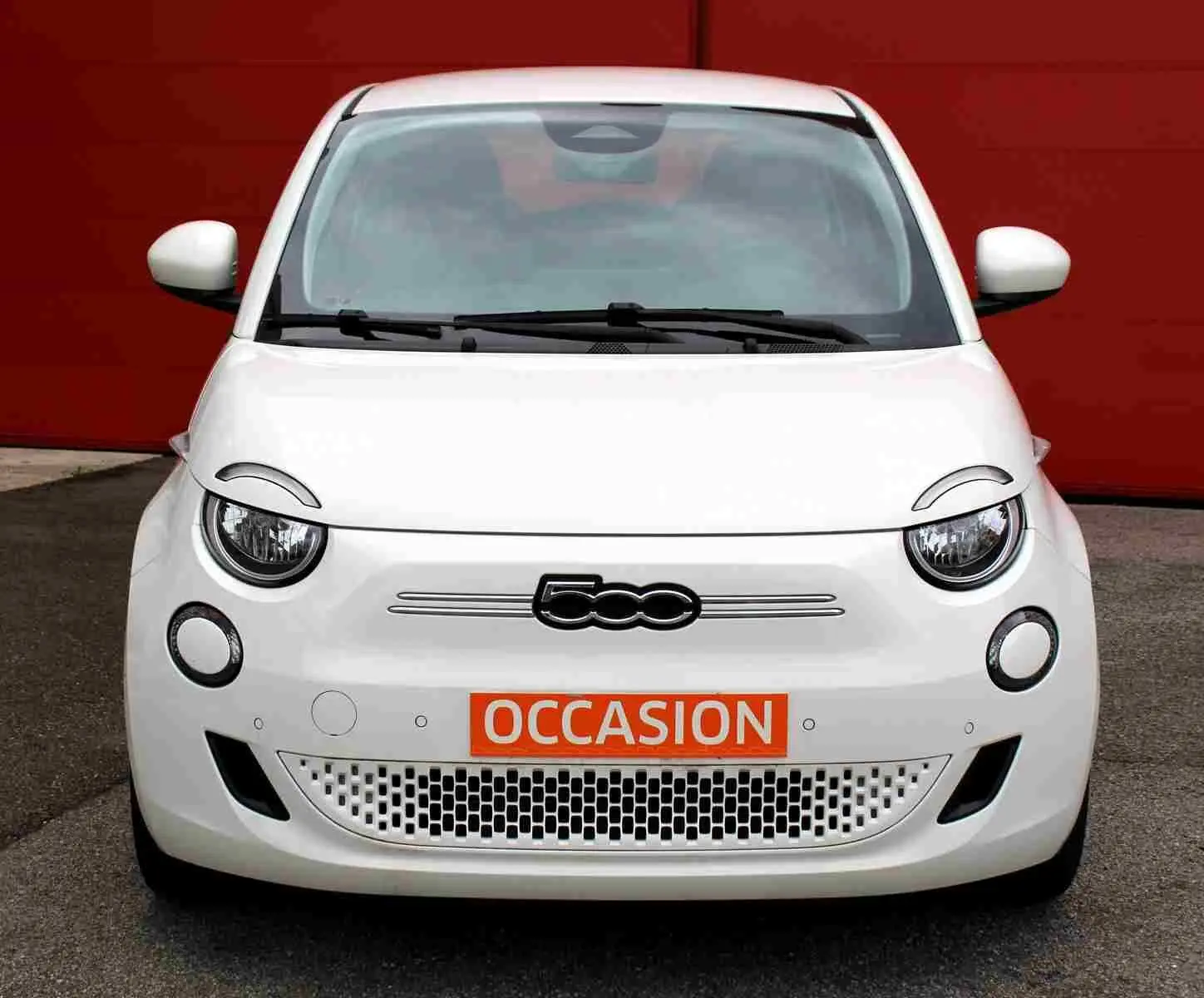 Vue avant d'une FIAT 500 électrique blanche avec logo distinctif et plaque orange "OCCASION" devant un mur rouge.