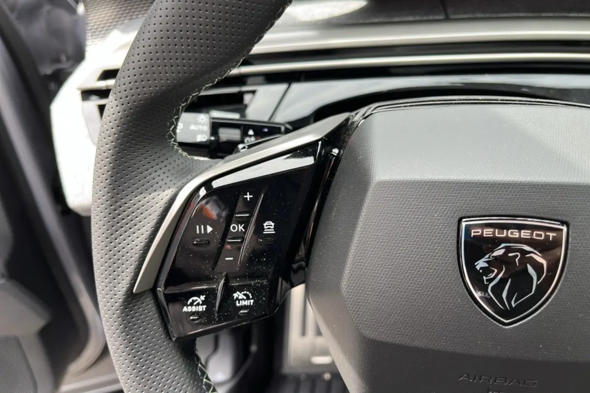 Gros plan sur le volant cuir perforé de la Peugeot 5008 Hybrid GT, avec commandes multifonctions et logo lion central.