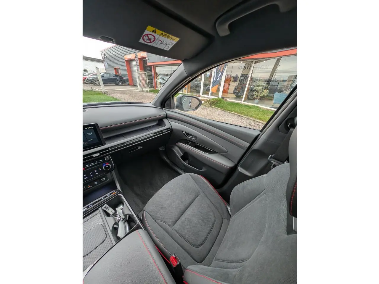 Vue intérieure côté passager du Hyundai Tucson gris, sellerie cuir/alcantara gris avec surpiqûres rouges et planche de bord noire.