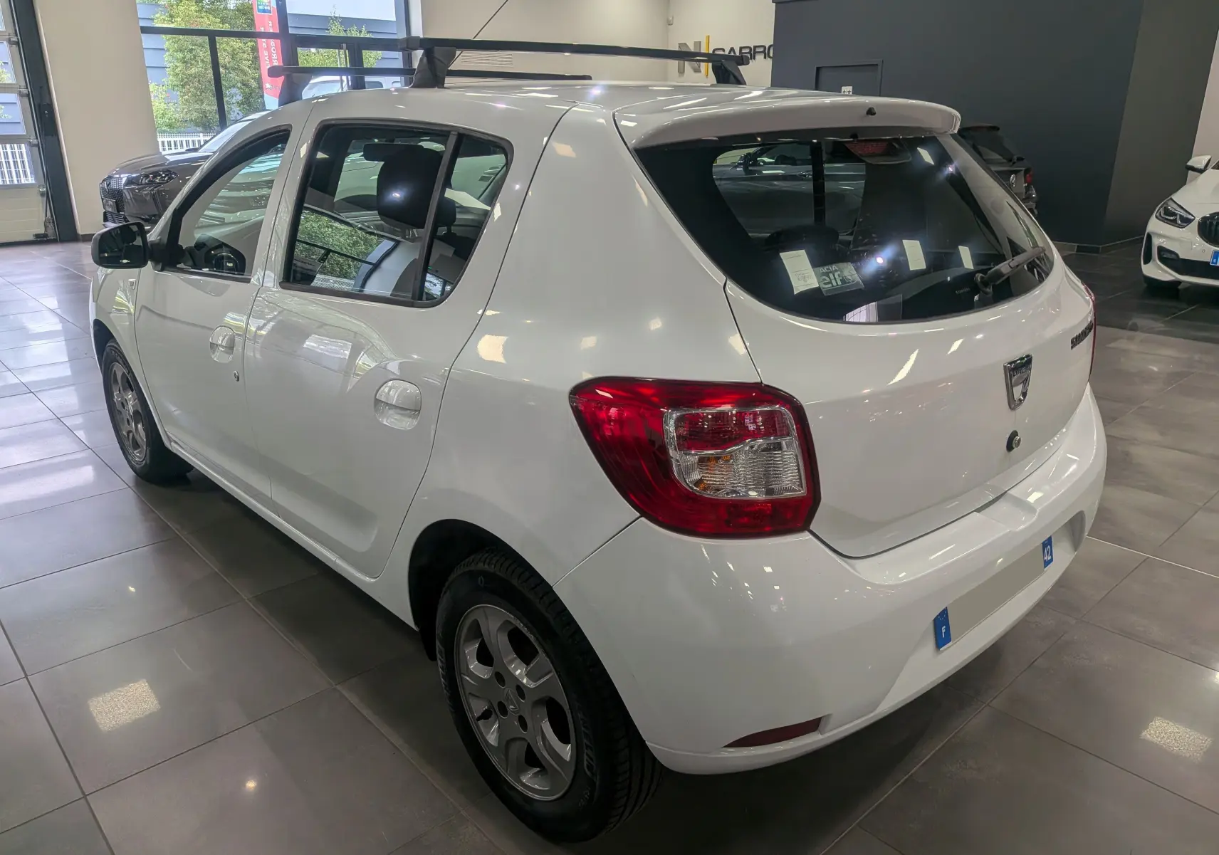 Vue 3/4 arrière droite d'une Dacia Sandero blanc glacier en intérieur, avec jantes alliage et vitres teintées.