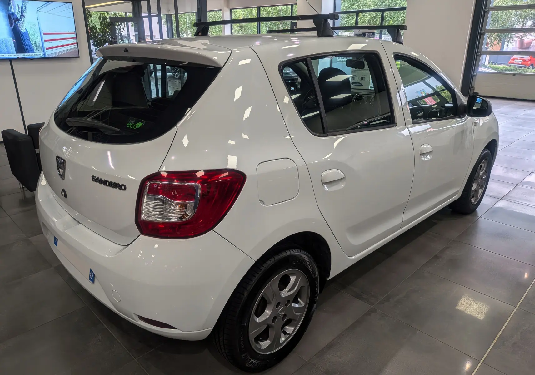 Vue 3/4 arrière droite d'une Dacia Sandero Blanc Glacier 2015 en showroom, avec jantes alliage et vitres teintées.