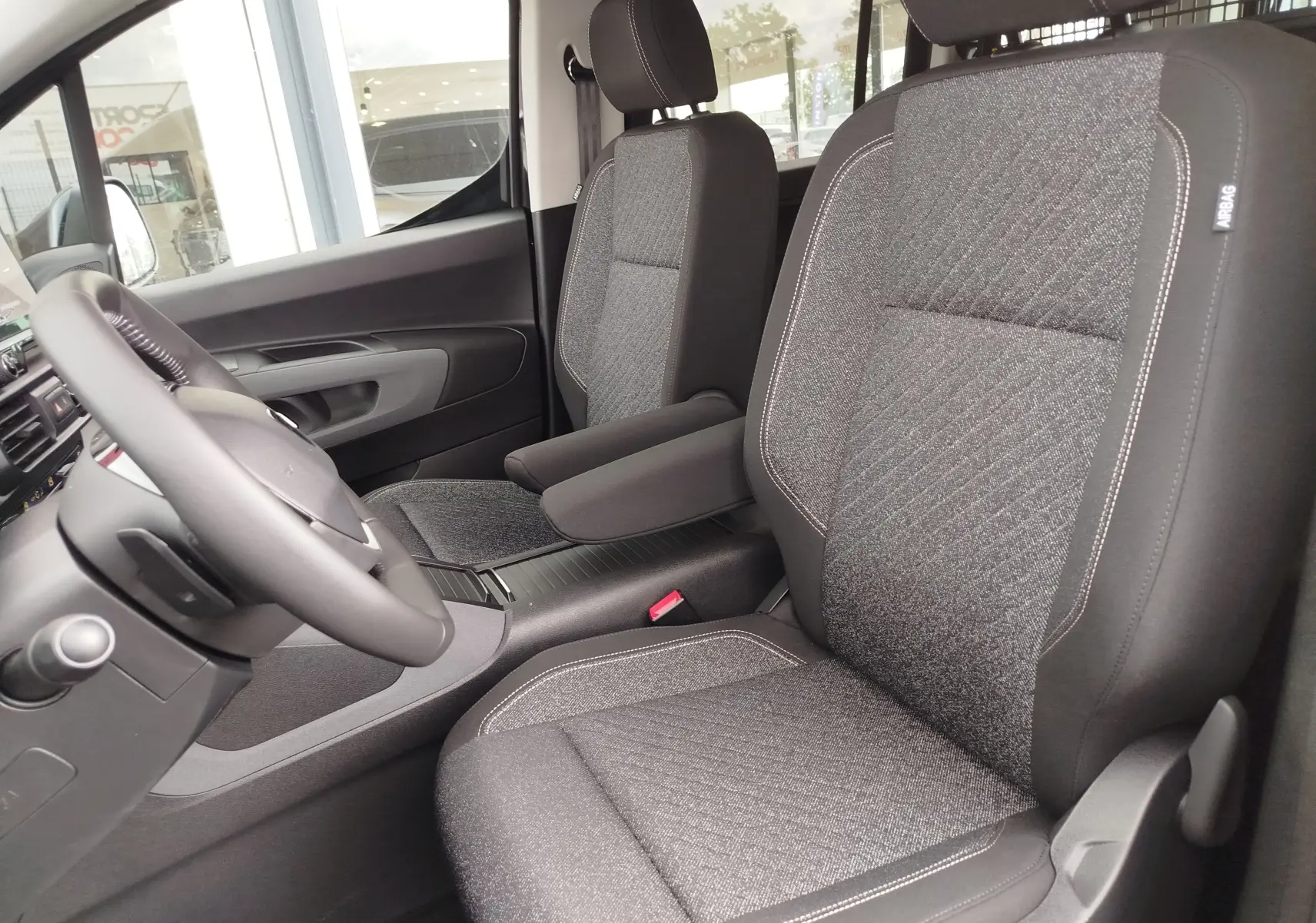 Vue intérieure côté conducteur du Peugeot Rifter 2026, sièges gris tissu avec coutures contrastées et accoudoir central.