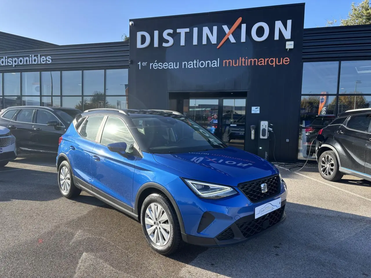 Vue 3/4 avant d'un SEAT Arona bleu saphir avec toit noir, stationné devant un bâtiment Distinxion.