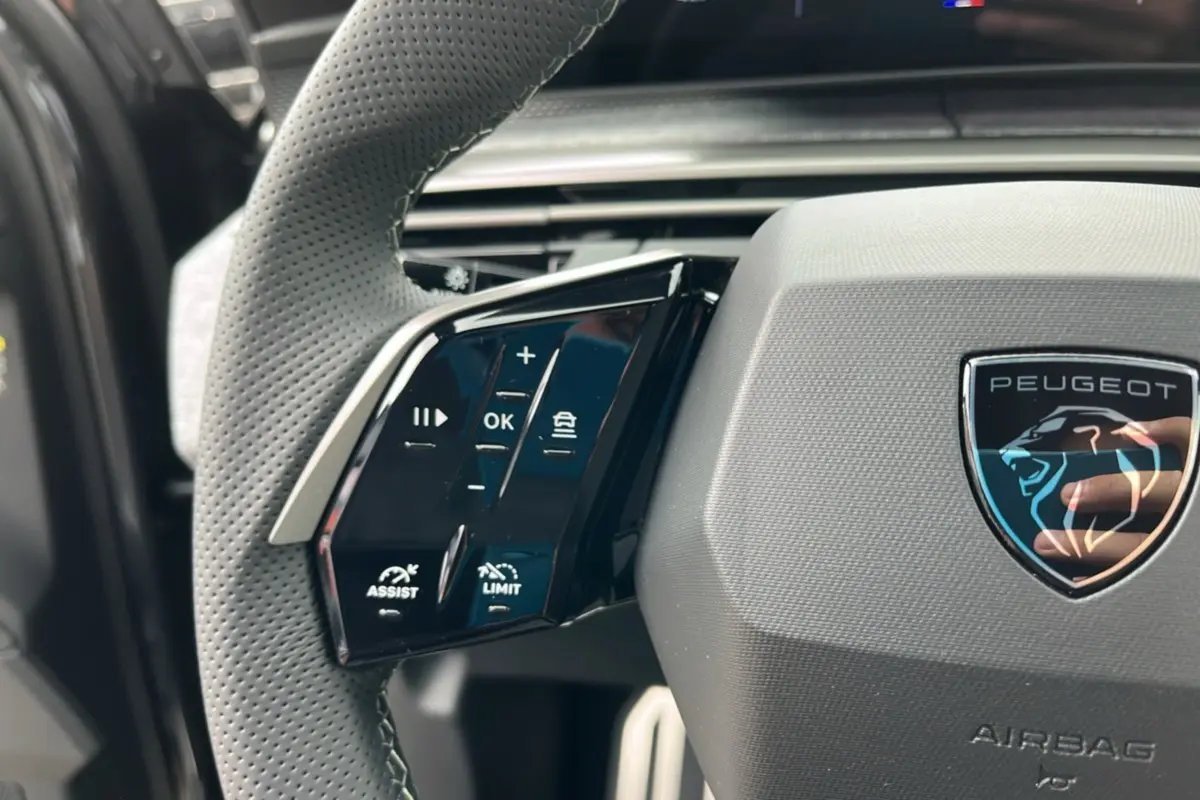 Gros plan sur le volant cuir perforé côté gauche du Peugeot 5008 Hybrid gris titane avec logo lion brillant.