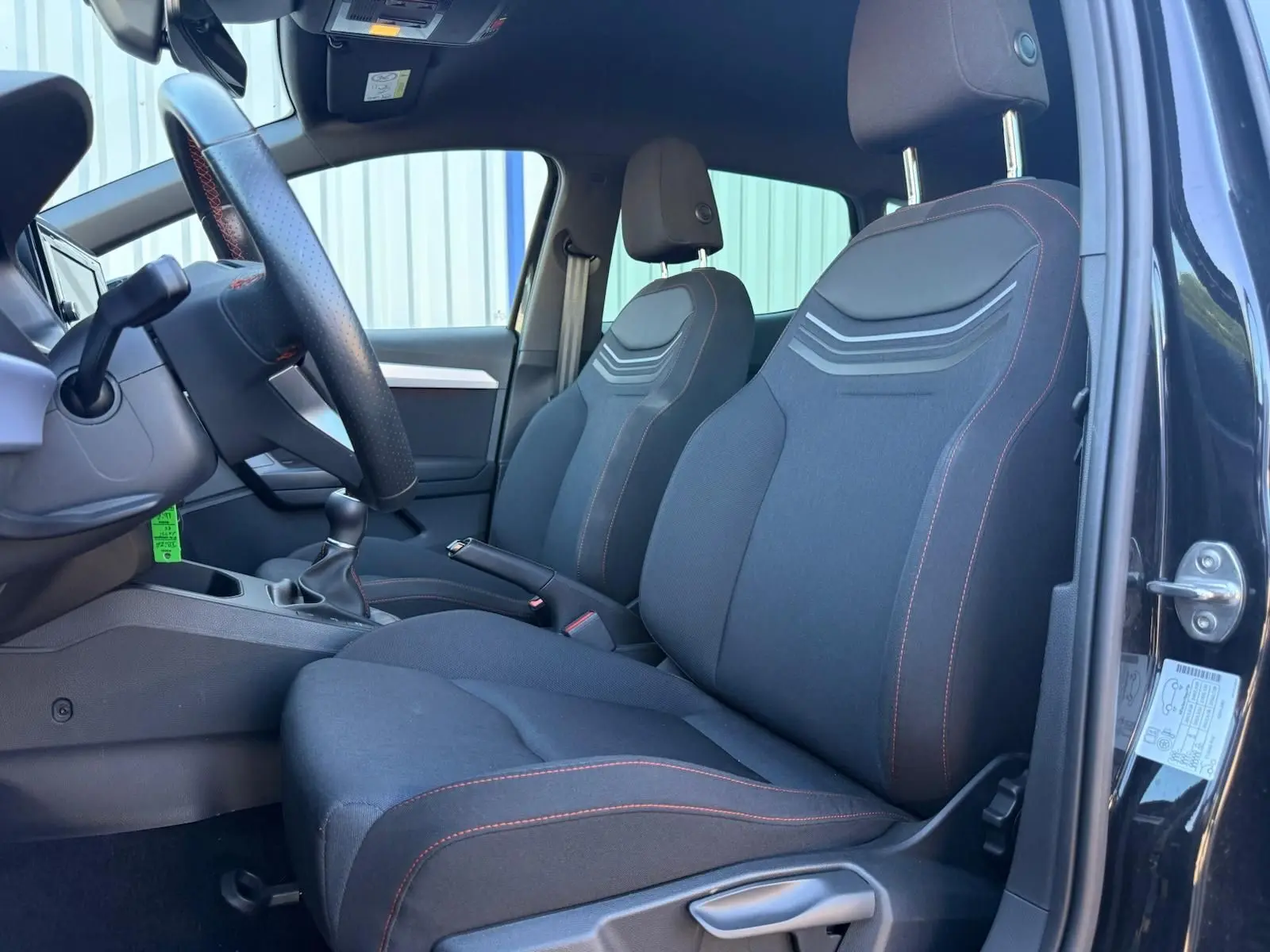 Vue intérieure côté conducteur de la SEAT IBIZA noire 2025, sièges tissu avec surpiqûres rouges et volant cuir.