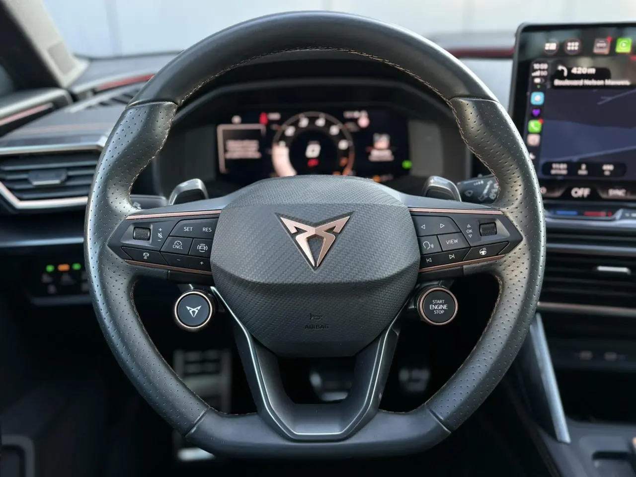 Vue rapprochée du volant cuir perforé noir avec logo CUPRA cuivré, tableau de bord digital et écran tactile en arrière-plan.