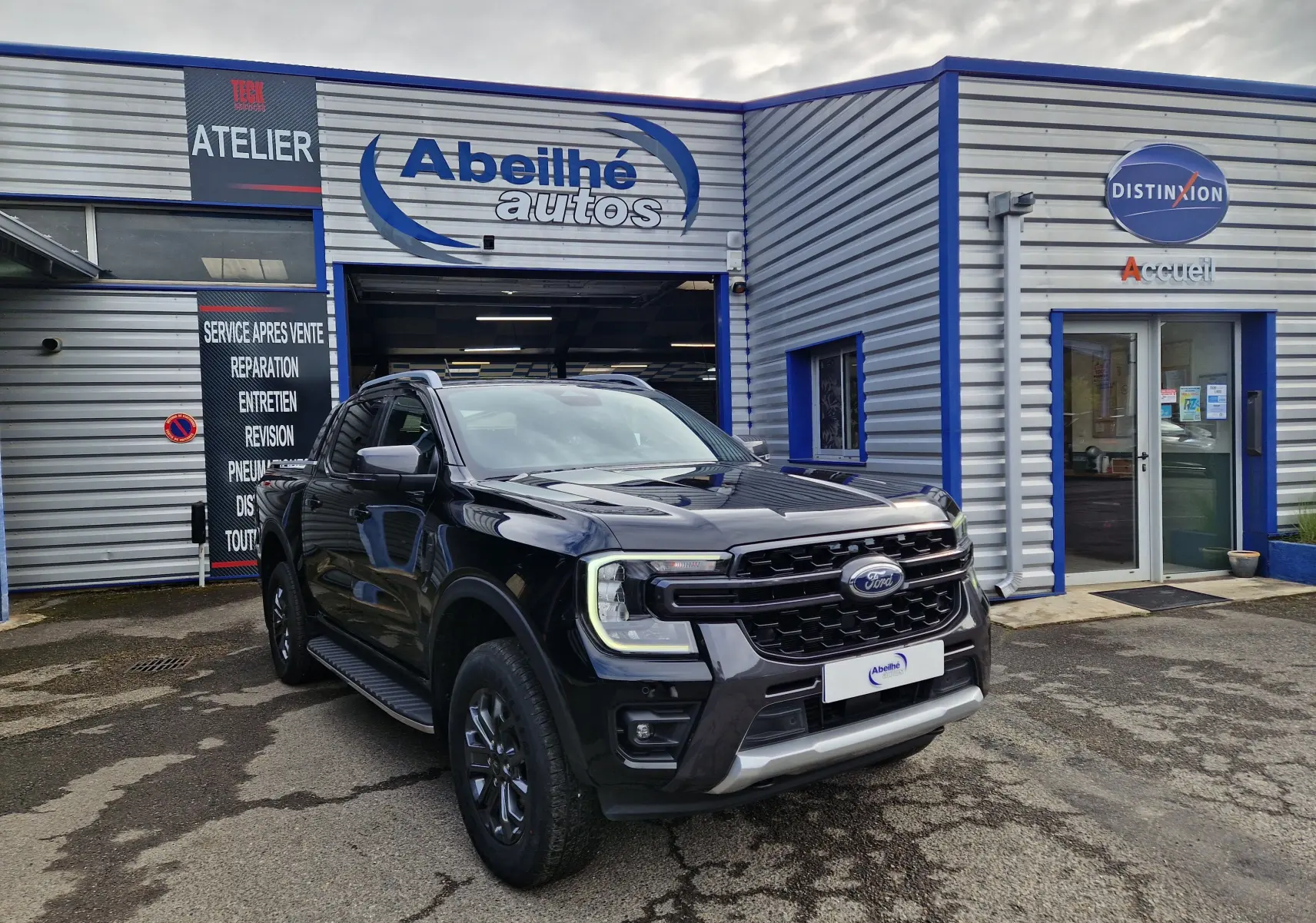 Ford Ranger Double Cabine noir en 3/4 avant droit, avec calandre imposante et marchepieds visibles devant un garage.