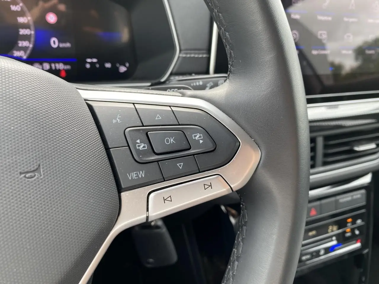 Gros plan sur les commandes gauche du volant cuir noir du Volkswagen T-Cross avec tableau de bord digital en arrière-plan.