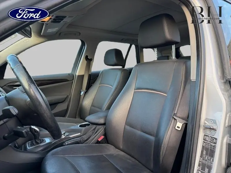 Intérieur avant d'une BMW X1 xDrive25dA 2014, sièges cuir noirs et volant multifonction visible côté conducteur.