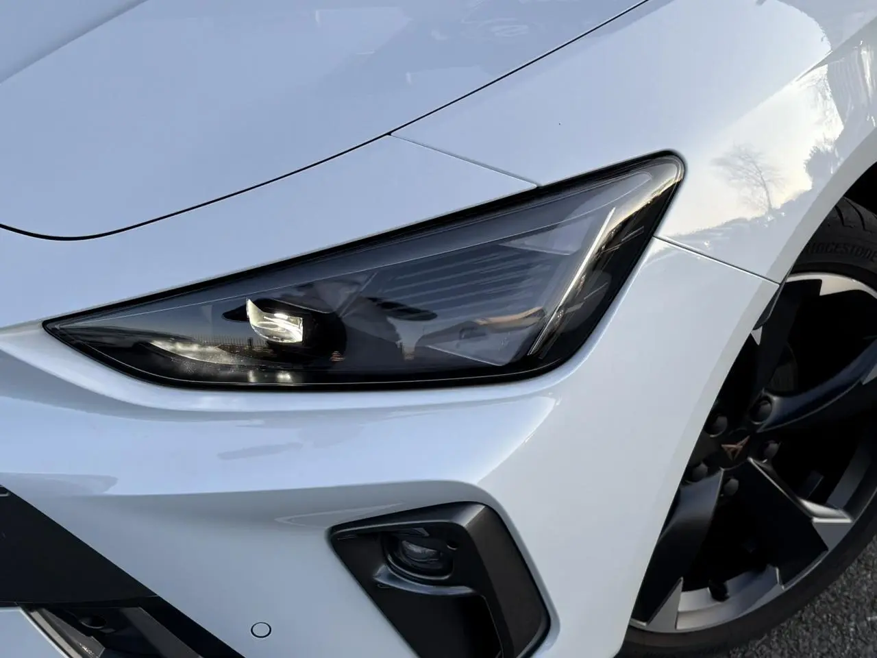 Gros plan sur le phare avant droit et la jante alliage noire d’une CUPRA Leon blanc neige.