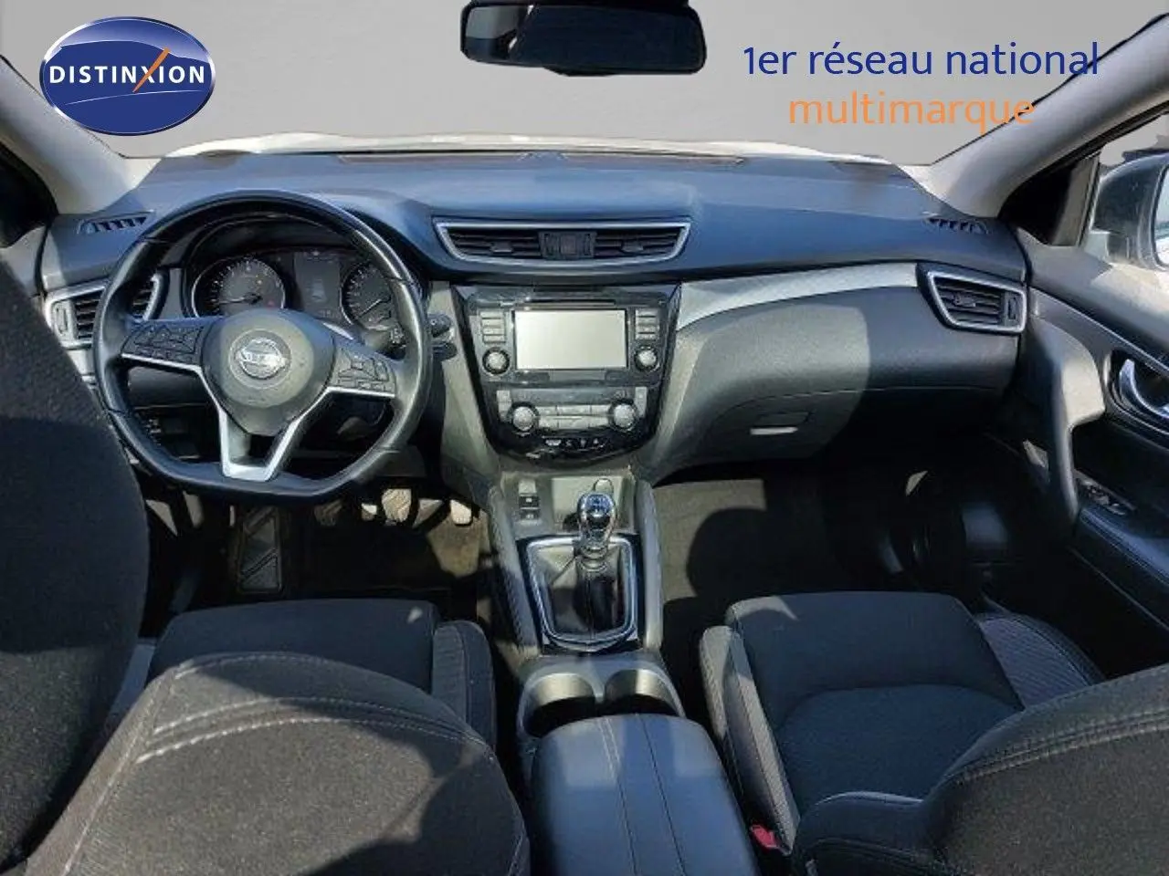 Vue intérieure du tableau de bord et console centrale du Nissan Qashqai blanc lunaire, avec volant et boîte manuelle visibles.