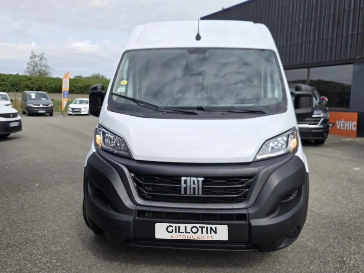 Vue frontale d'un FIAT Ducato Fourgon blanc avec calandre noire et phares allumés, sur parking extérieur.