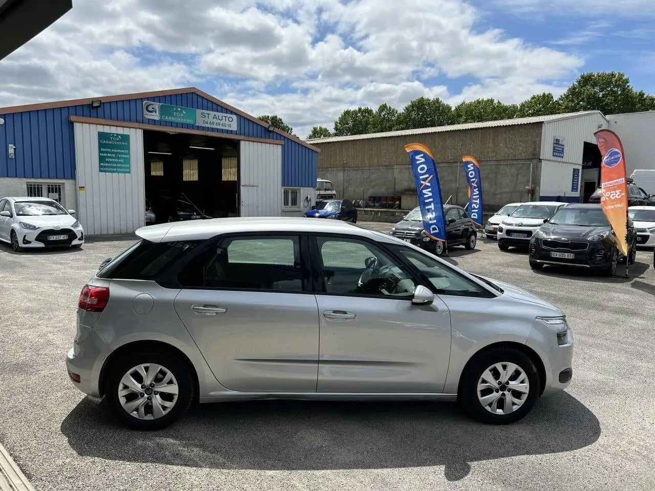 Profil côté droit d'une Citroën C4 Picasso gris aluminum 2016 stationnée devant un garage, sous un ciel partiellement nuageux.