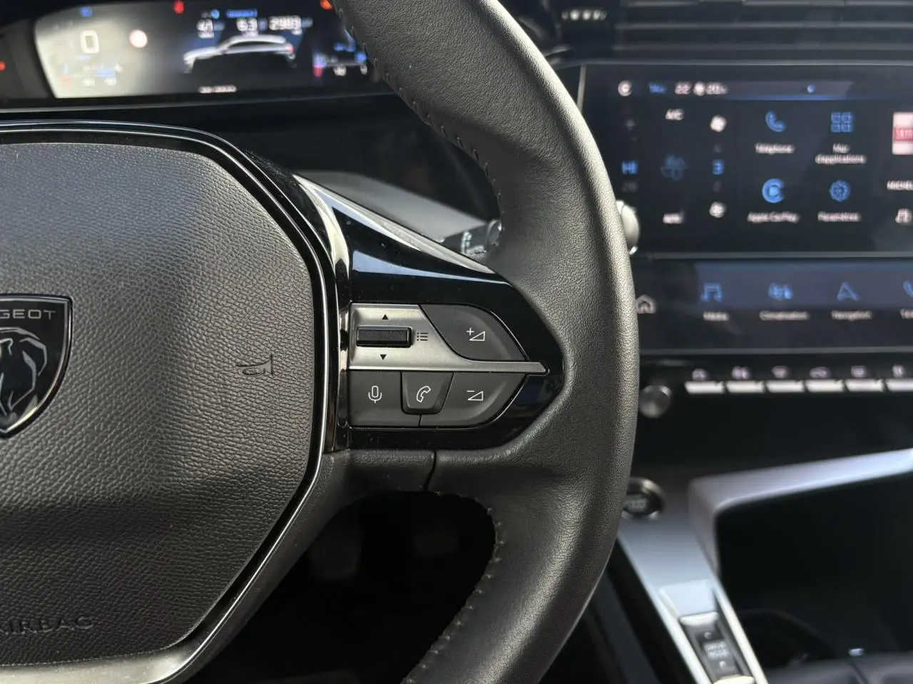 Gros plan sur le volant cuir noir de la Peugeot 308 2024 avec commandes vocales et écran tactile en arrière-plan flou.