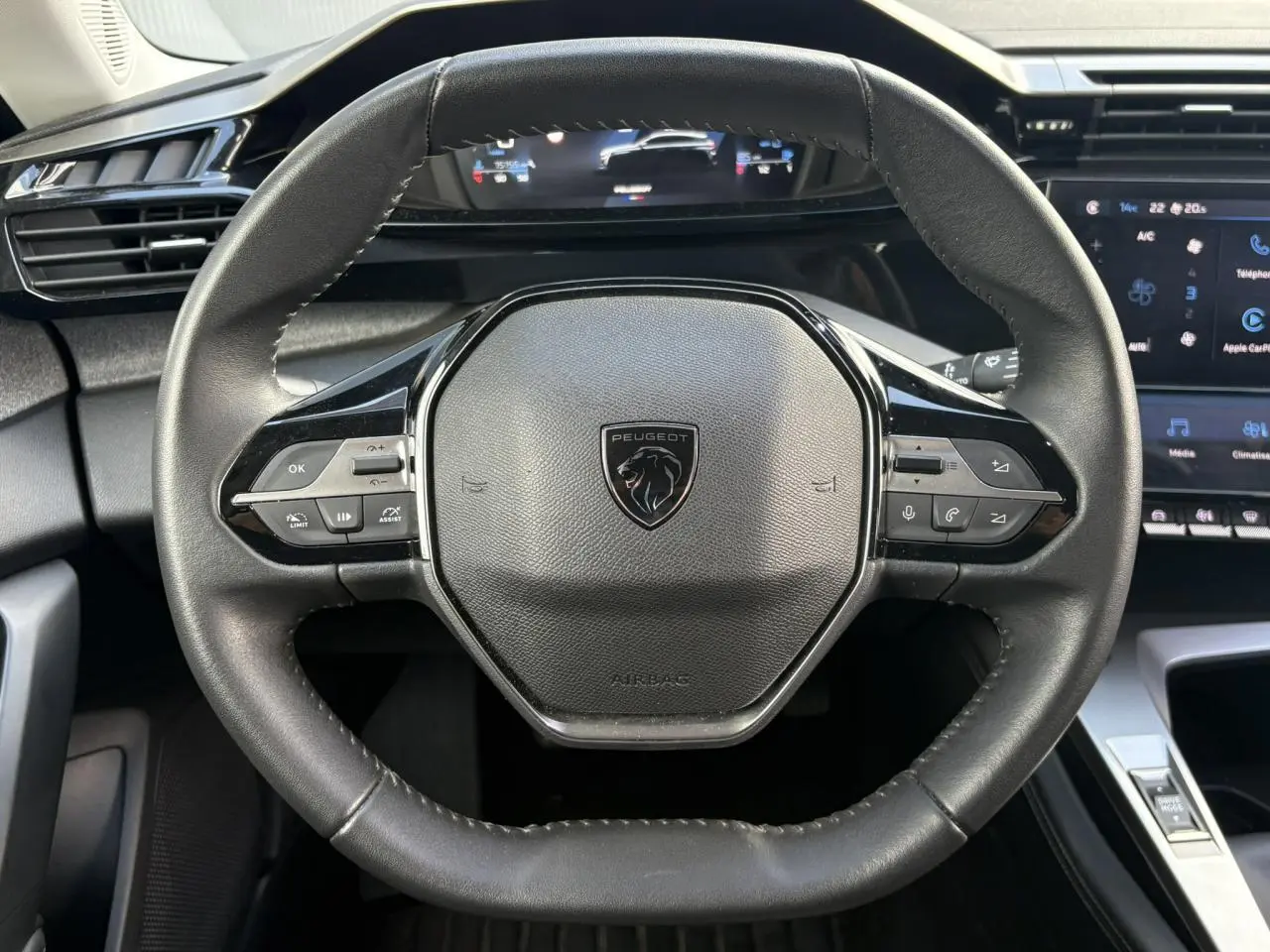 Vue rapprochée du volant cuir noir au design hexagonal de la Peugeot 308 2024 avec tableau de bord numérique en arrière-plan.