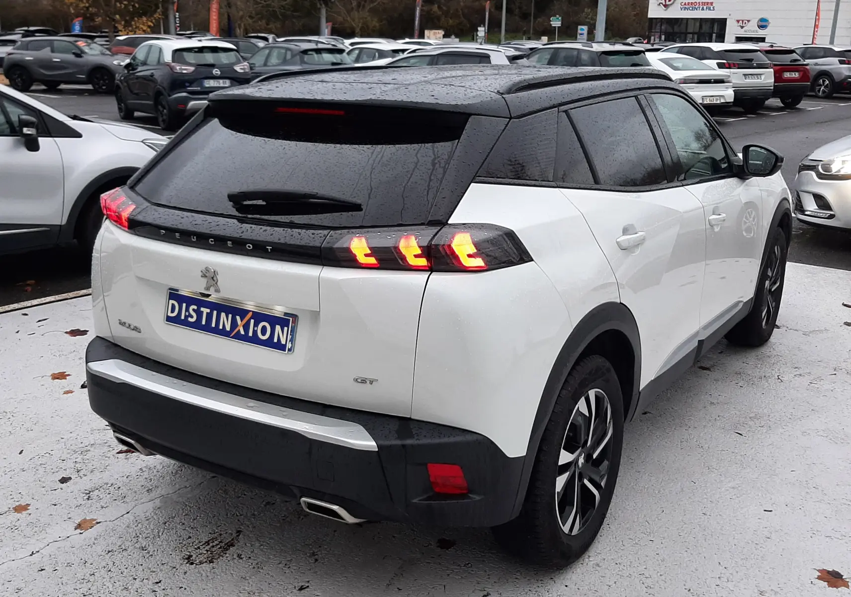 Vue 3/4 arrière droite d'un Peugeot 2008 blanc nacré avec toit noir, feux arrière allumés et attelage visible.
