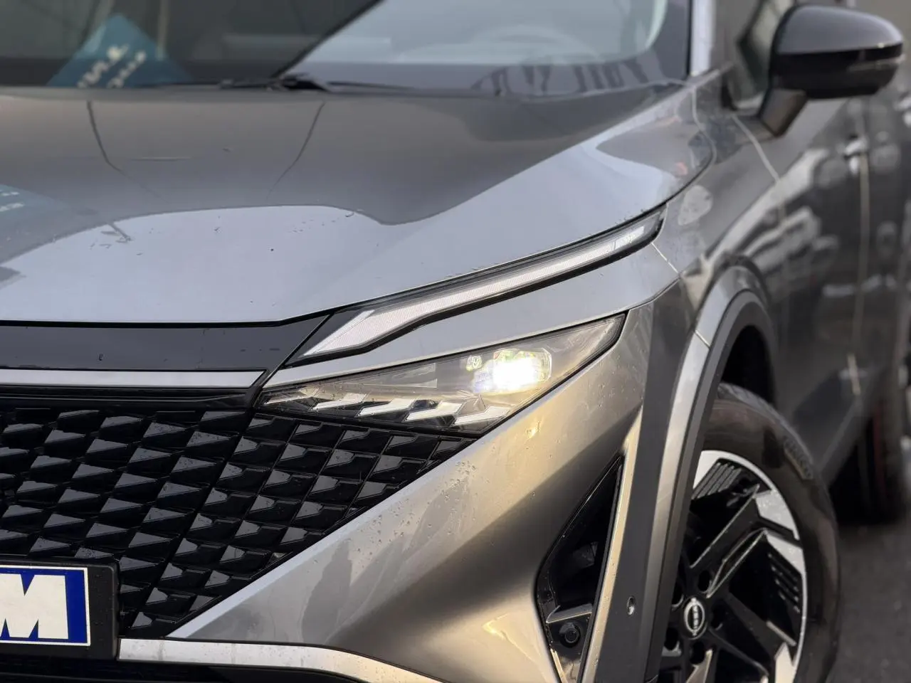 Gros plan sur l'avant droit du Nissan Qashqai 2025 gris Squale, avec phares allumés et jante alliage distinctive.