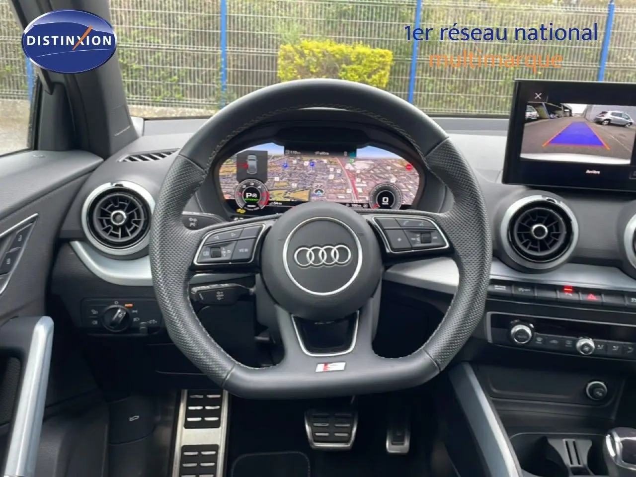 Intérieur Audi Q2 2025 vu du poste de conduite, volant cuir S-Line, tableau de bord numérique et écran caméra de recul.