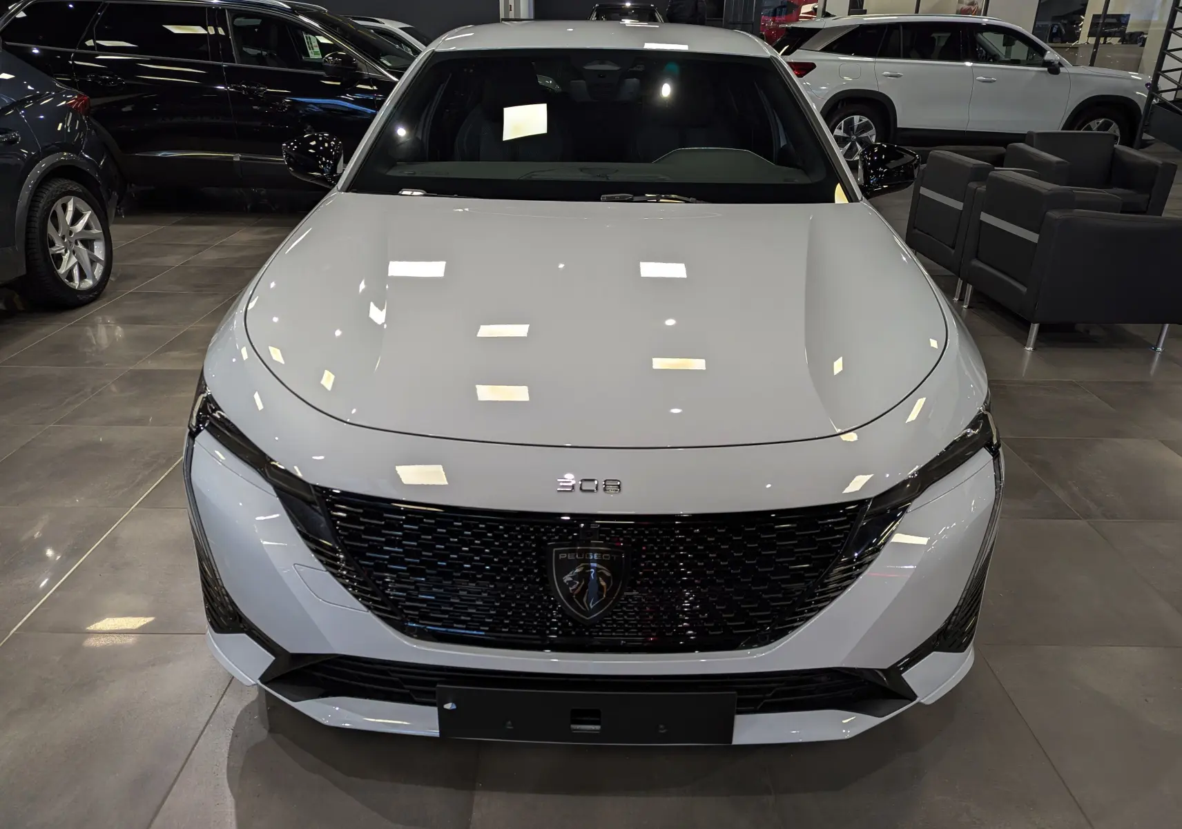 Vue frontale d'une Peugeot 308 2024 blanche Okénite avec calandre noire et logo lion au centre, en showroom.