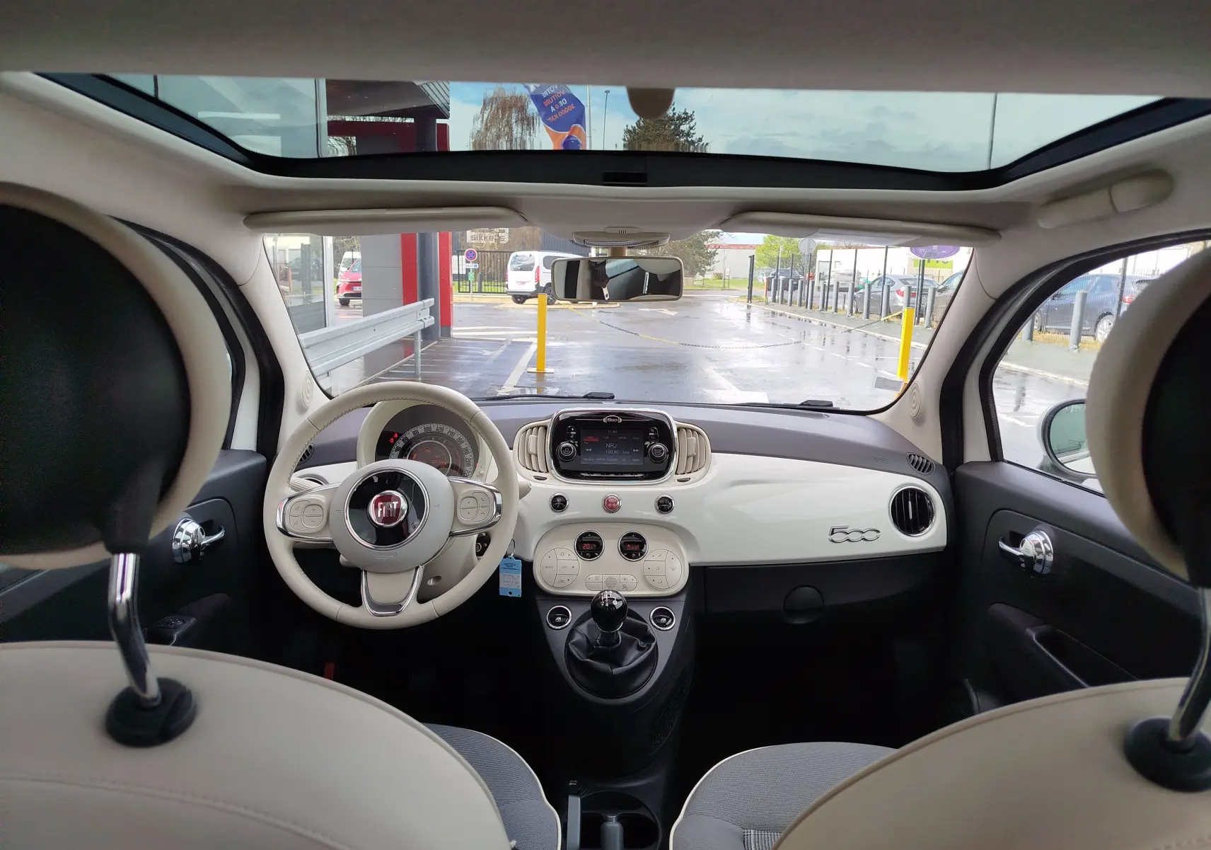Vue intérieure avant de la Fiat 500 blanche avec tableau de bord épuré, volant beige et toit panoramique ouvert.