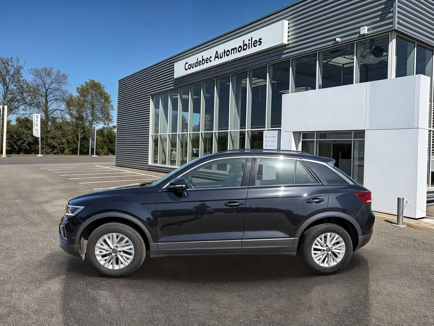 Profil côté gauche d’un Volkswagen T-Roc noir 2025 garé devant un bâtiment moderne de concession automobile.