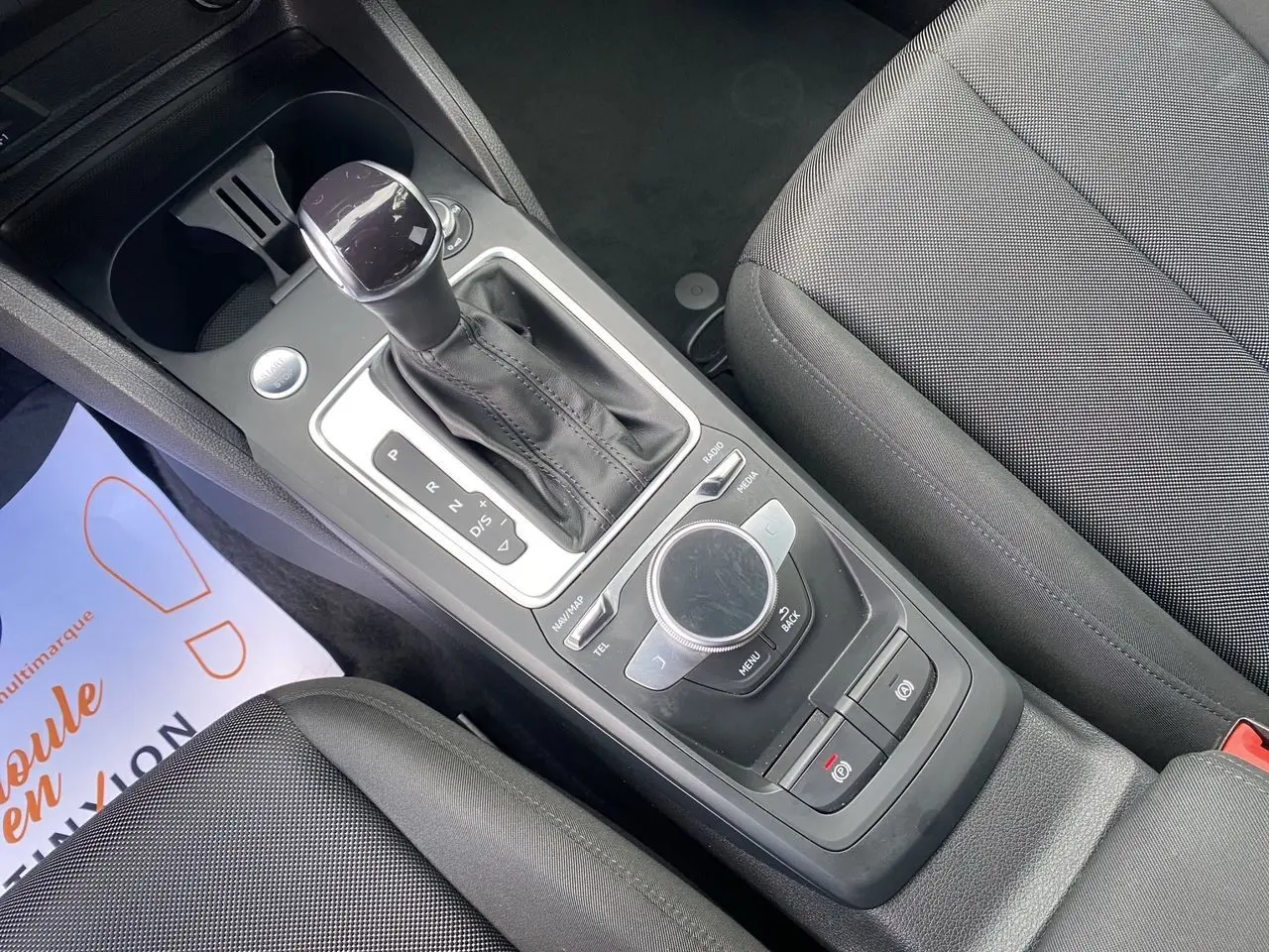 Vue rapprochée de la console centrale noire de l'Audi Q2 2023, avec levier de vitesse automatique et molette de contrôle multimédia.