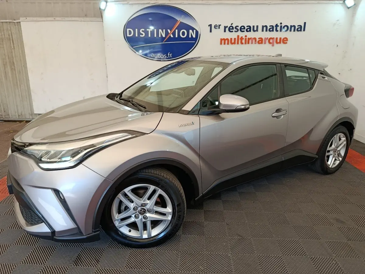 Vue 3/4 avant droit d’un Toyota C-HR 2.0 Hybride gris clair, avec jantes alliage et détails hybrides visibles sur l’aile.