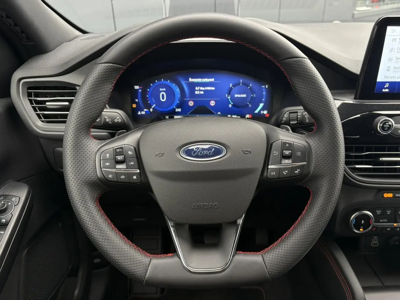 Vue rapprochée du volant perforé noir avec surpiqûres rouges du Ford Kuga 2023, tableau de bord numérique en arrière-plan.