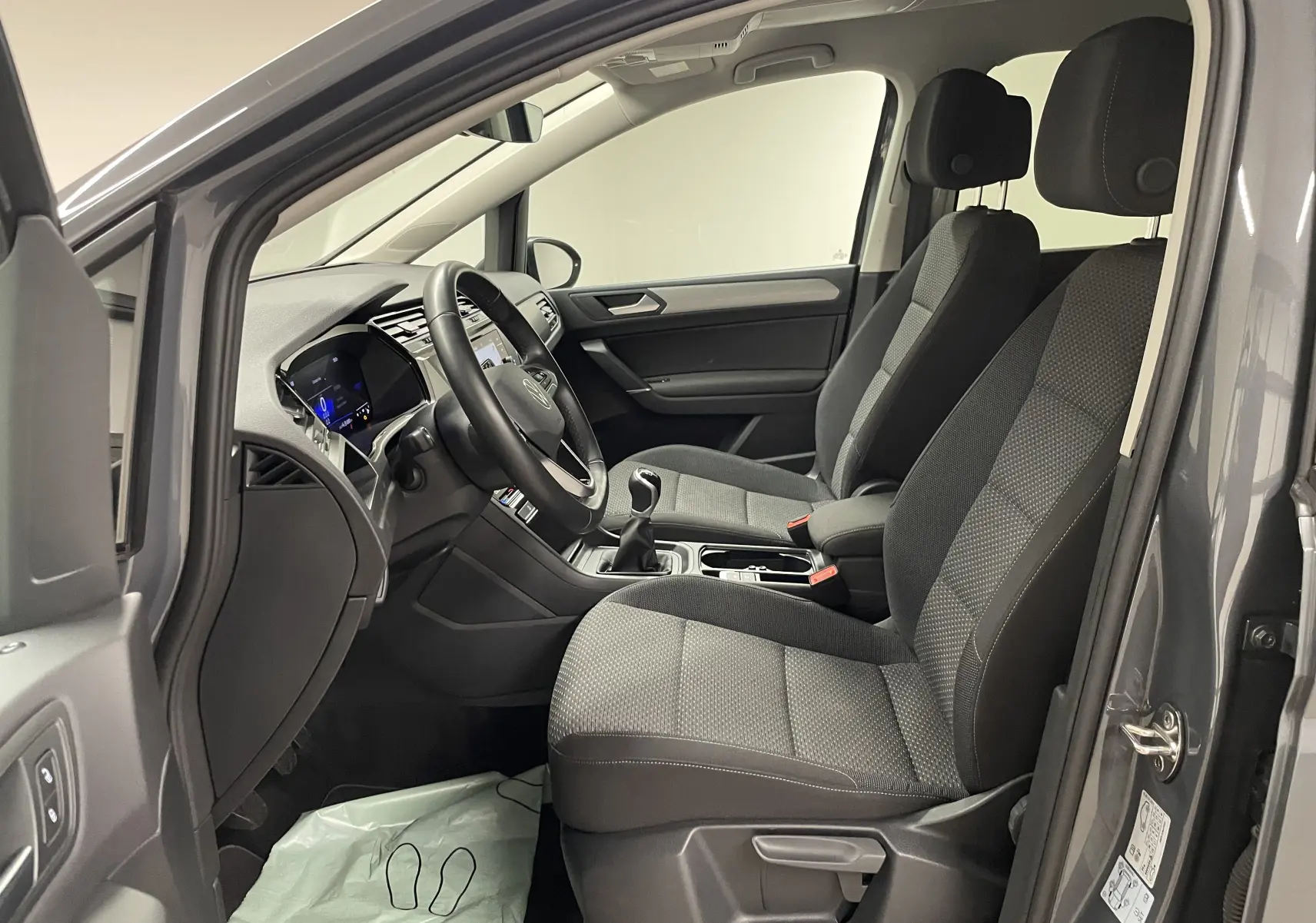 Vue intérieure côté conducteur du Volkswagen Touran gris Dauphin 2024, sièges tissu et tableau de bord moderne avec boîte manuelle.