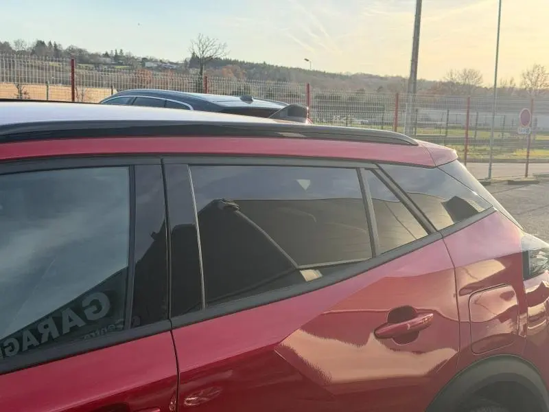 Vue latérale droite d'un Peugeot 2008 rouge Elixir avec vitres arrière teintées et barres de toit visibles.