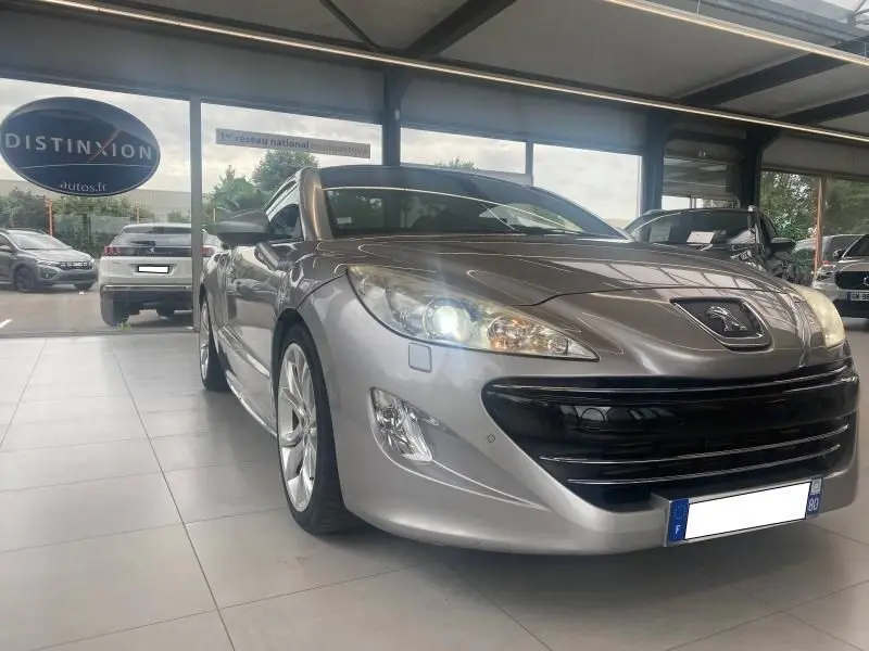 Peugeot RCZ gris vu en 3/4 avant droit avec phares allumés dans un showroom lumineux.