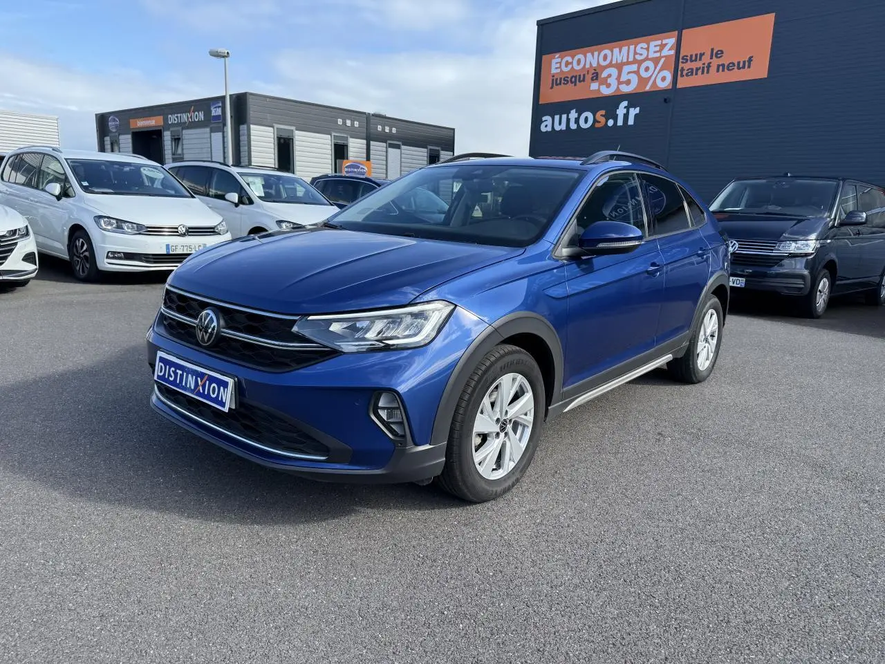 Volkswagen Taigo bleu récif métallisé vu en 3/4 avant droit sur parking devant un panneau publicitaire orange.