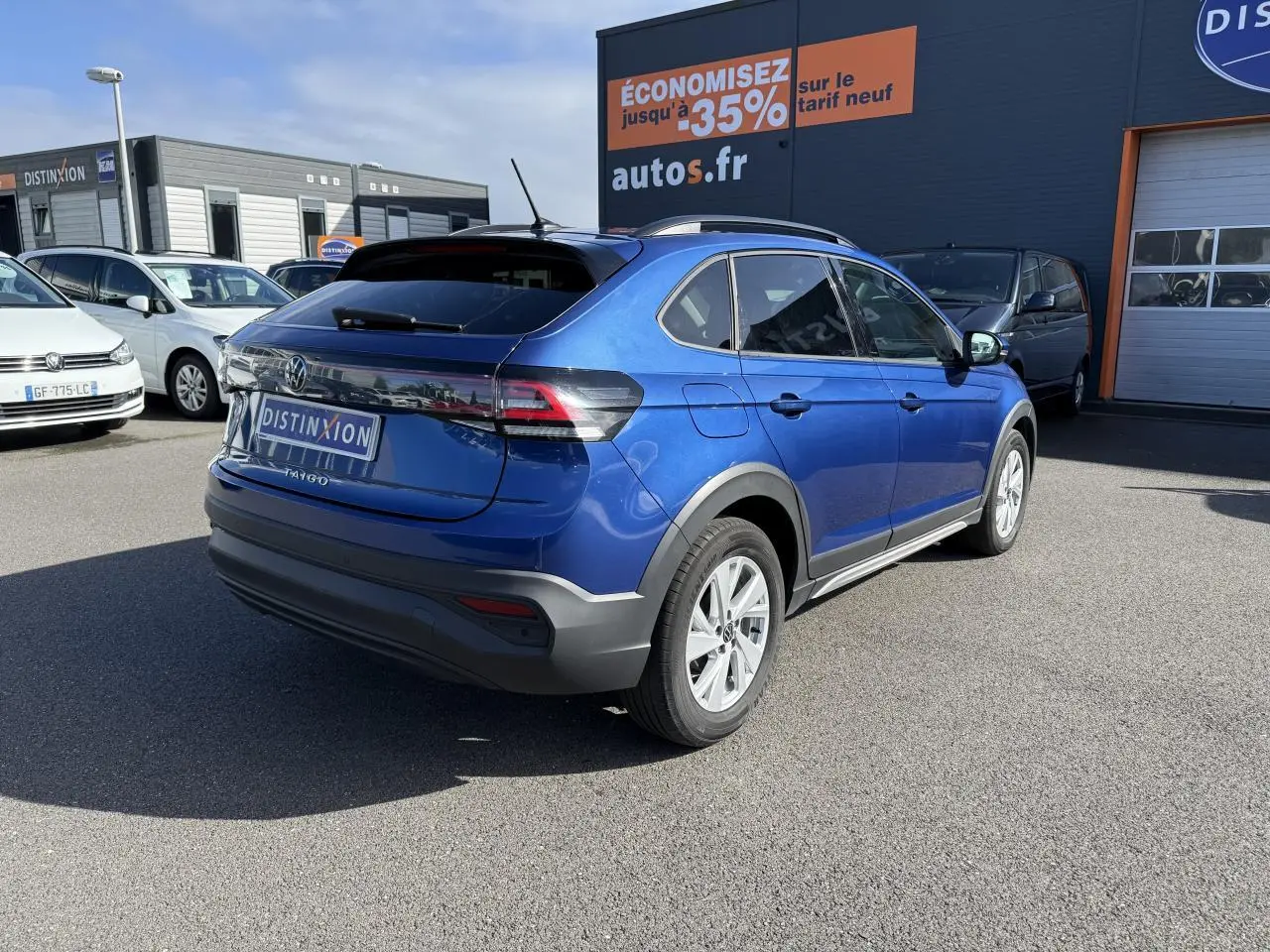 Volkswagen Taigo bleu récif métallisé vu en 3/4 arrière droit sur parking devant un bâtiment commercial.