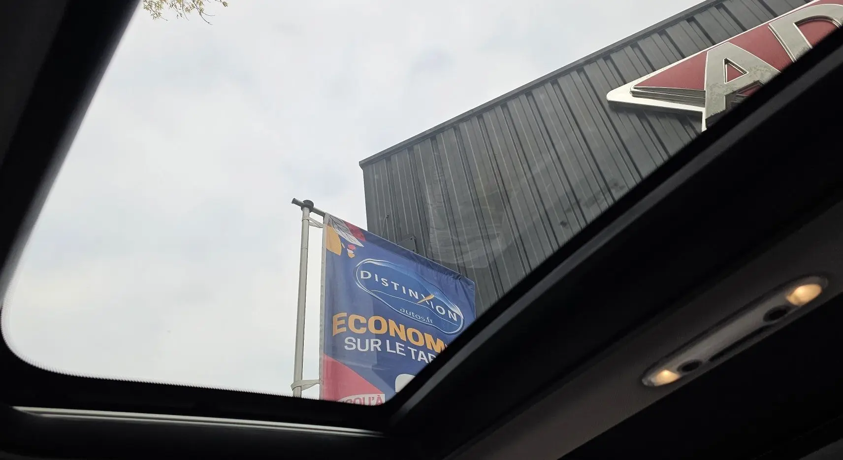 Vue partielle par le toit ouvrant d'un MINI Cooper S Clubman grise 2022, avec ciel nuageux et enseigne de concession visible.