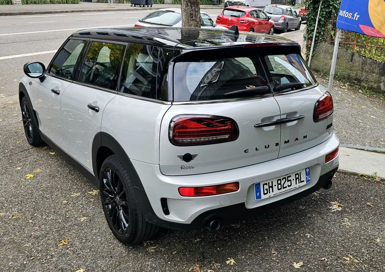 Vue 3/4 arrière droite d'une MINI Clubman grise avec toit noir et jantes noires, stationnée sur un parking urbain.