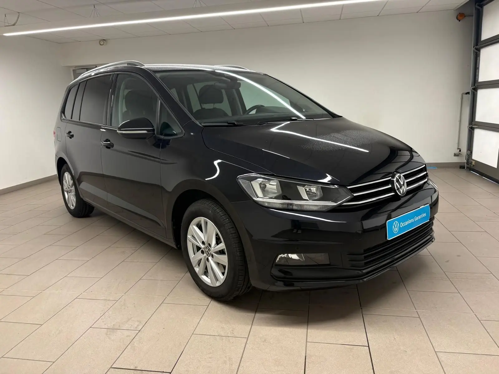 Volkswagen Touran noir intense nacré vu en 3/4 avant droit dans un garage lumineux avec jantes argentées.