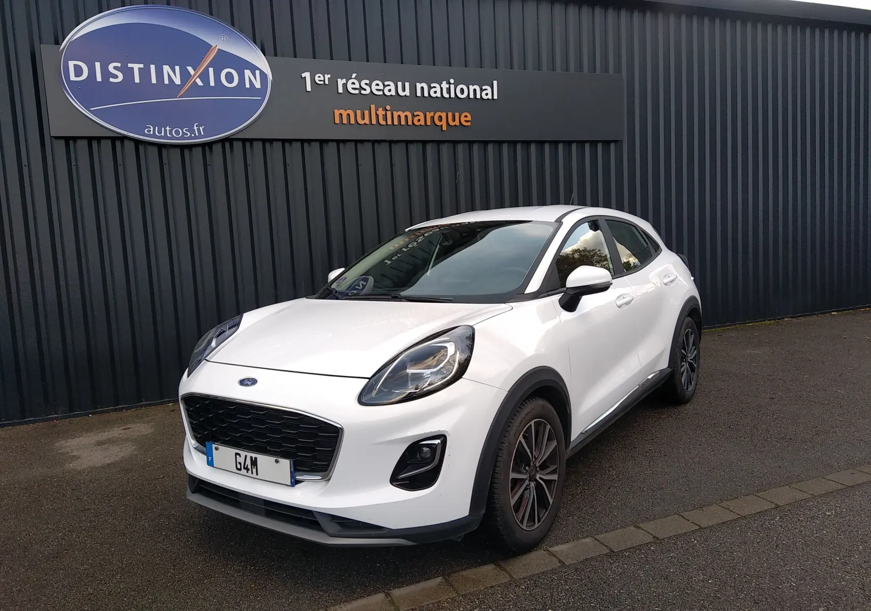 Ford Puma blanc en 3/4 avant droit, avec calandre noire et jantes foncées, stationné devant un mur gris.