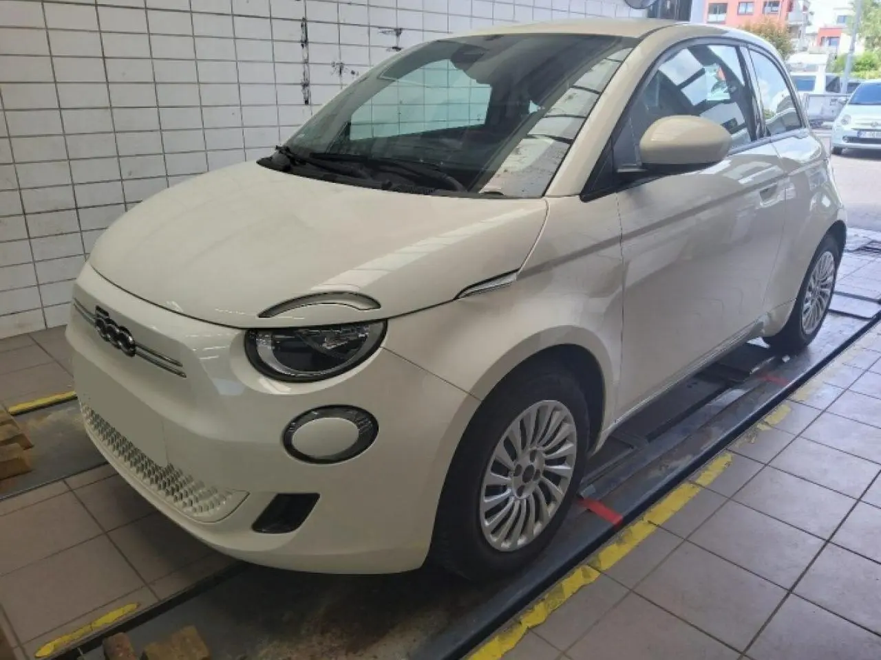 Vue 3/4 avant droite d'une Fiat 500 électrique blanche stationnée dans un garage avec phares ronds distinctifs.