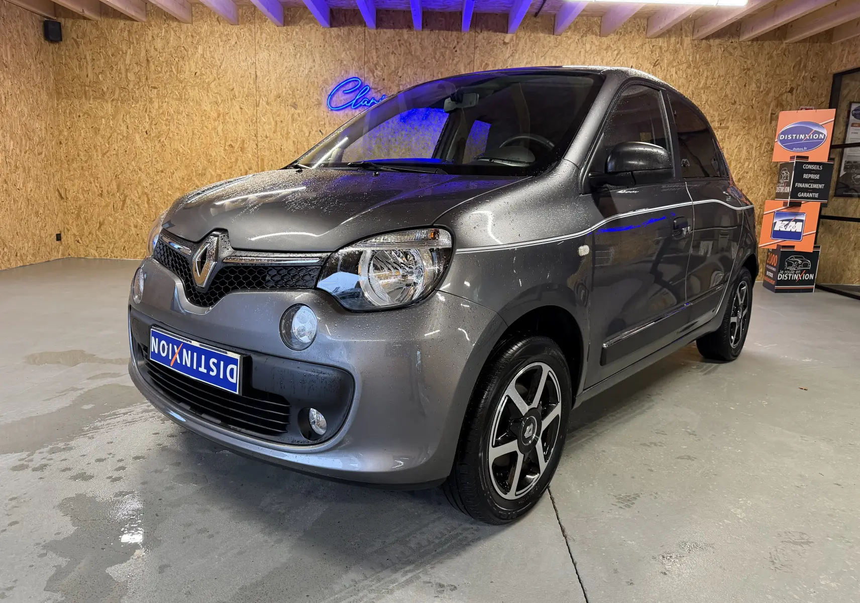 Renault Twingo III gris en 3/4 avant droit, avec jantes noires et phares halogènes visibles en intérieur showroom.