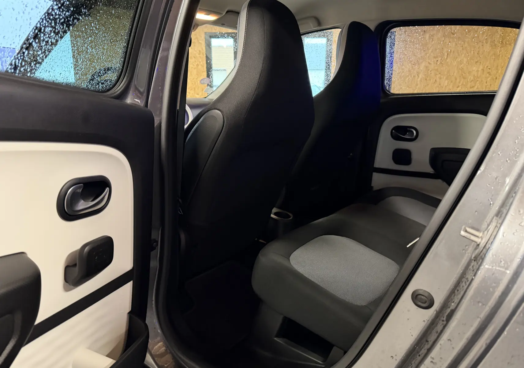 Vue intérieure côté droit de la Renault Twingo III gris, montrant les sièges arrière et la porte ouverte sous la pluie.